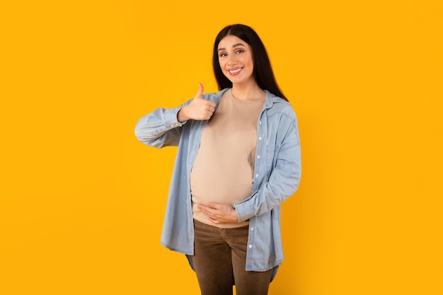 Heureuse femme attendant un bébé touchant le ventre de femme enceinte et gesticulant les pouces vers le haut souriant à l'arrière-plan jaune de la caméra