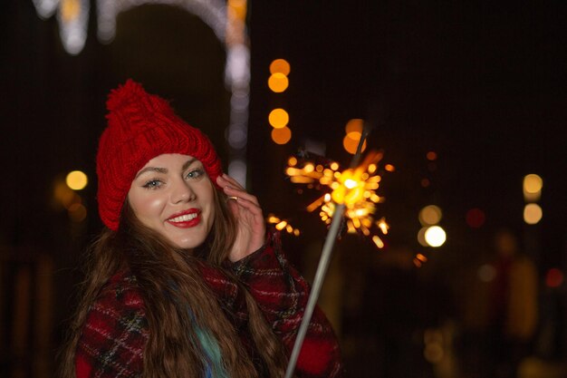 Heureuse femme au chapeau rouge s'amusant avec des lumières étincelantes en plein air le soir. Espace libre