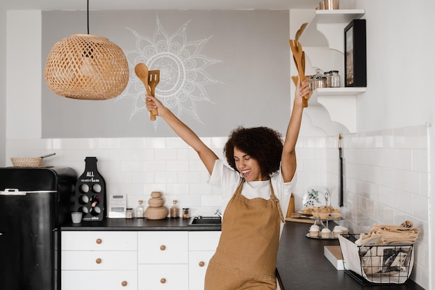 Heureuse femme au foyer africaine en tablier chantant et dansant sur la cuisine avant de cuisiner le petit déjeuner Cuisinière afro-américaine dansant avec des spatules pour cuisiner et s'amuser