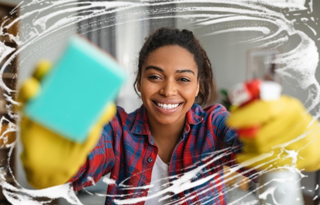 Photo heureuse femme au foyer afro-américaine du millénaire dans des gants en caoutchouc avec un spray et une éponge lave le miroir avec de la mousse