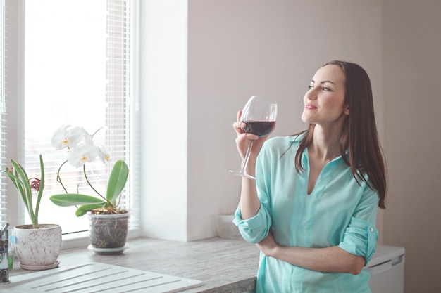 Heureuse femme au foyer dans la cuisine avec un verre de vin rouge à la recherche dans la fenêtre