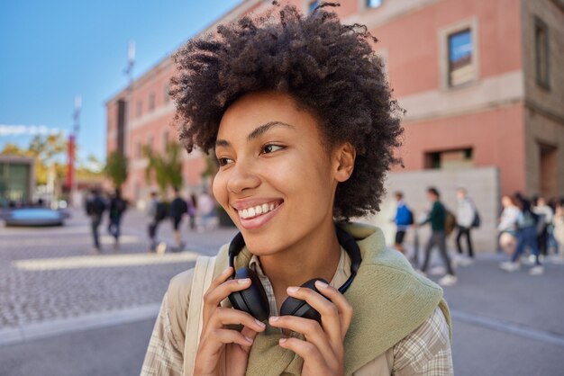 Heureuse femme aux cheveux bouclés avec un casque électronique autour du cou regarde joyeusement loin explore la ville en vacances vêtue de vêtements décontractés pose dans la rue sur fond flou Concept de style de vie