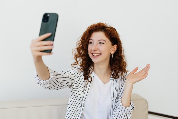 Heureuse femme aux cheveux roux bouclés se reposant à la maison sur un canapé en cuir parlant en train de discuter via un appel vidéo à l'aide d'une application sur un smartphone a une conversation avec la famille saluant des amis en agitant la main