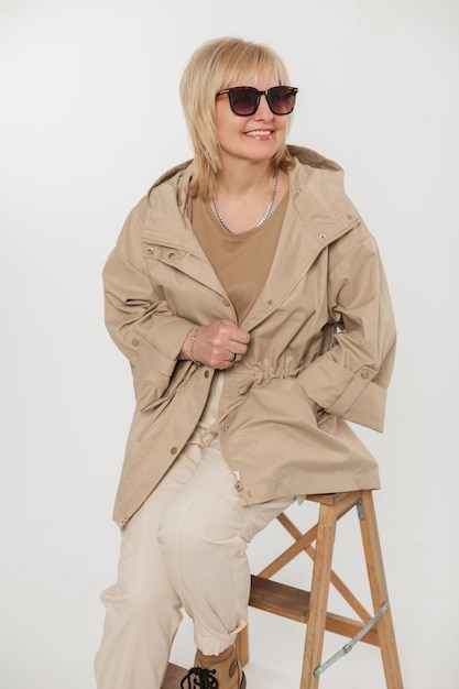 Heureuse femme blonde âgée de mode avec un sourire avec des lunettes de soleil vintage dans des vêtements beiges à la mode avec une veste est assise sur une chaise en bois sur fond blanc