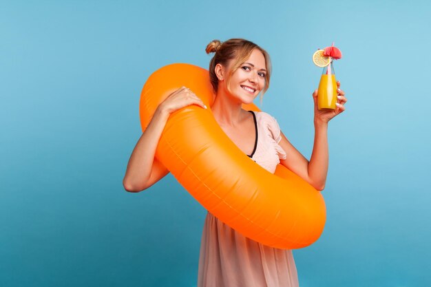 Heureuse femme blonde avec un anneau en caoutchouc orange levant la main avec un cocktail et regardant directement la caméra avec un sourire charmant, portant une robe d'été. Studio intérieur tourné isolé sur fond bleu.