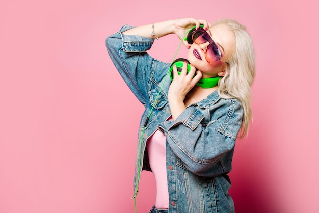 Heureuse femme blonde élégante dans des verres roses avec de gros écouteurs écoutant de la musique et dansant sur fond coloré