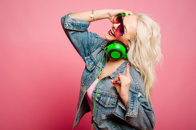 Heureuse femme blonde élégante dans des verres roses avec de gros écouteurs écoutant de la musique et dansant sur fond coloré