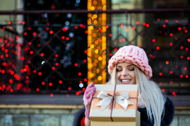 Heureuse femme blonde portant une casquette tricotée et un manteau chaud, ouvrant une boîte-cadeau à l'arrière-plan des lumières de la ville. Espace pour le texte