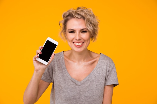 Heureuse femme blonde en pull montrant un écran de smartphone vierge et pointant dessus tout en regardant la caméra avec la bouche ouverte sur fond jaune