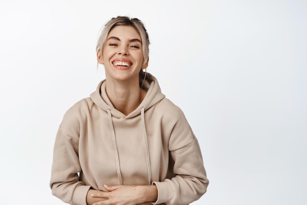 Heureuse femme blonde en riant et en regardant quelque chose de drôle tenant les mains sur le ventre et souriant debout sur fond blanc