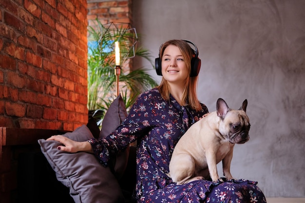 Heureuse femme blonde en robe assise avec son joli carlin sur un canapé fait main et écoutant de la musique dans la chambre avec intérieur loft.