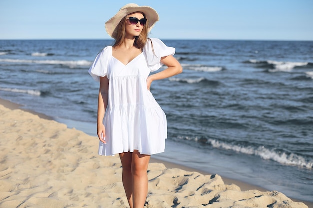 Heureuse femme blonde souriante pose sur la plage de l'océan avec des lunettes de soleil et un chapeau.