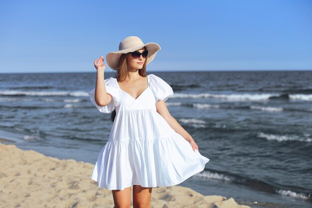 Heureuse femme blonde souriante pose sur la plage de l'océan avec des lunettes de soleil et un chapeau.