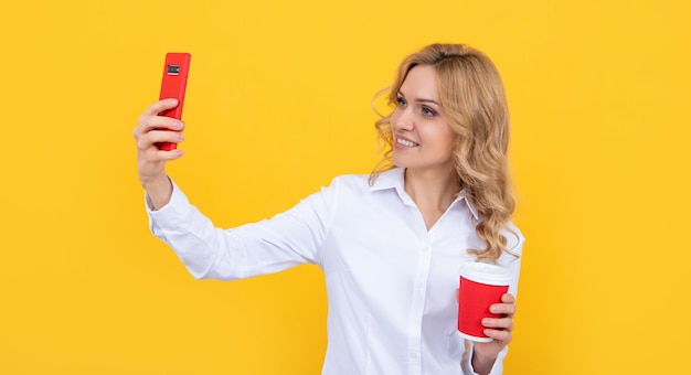 Heureuse femme blonde avec une tasse de café faisant selfie au téléphone sur fond jaune