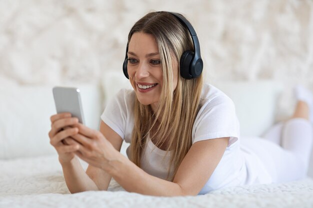 Heureuse femme blonde utilisant un téléphone portable et un casque à l'intérieur de la chambre