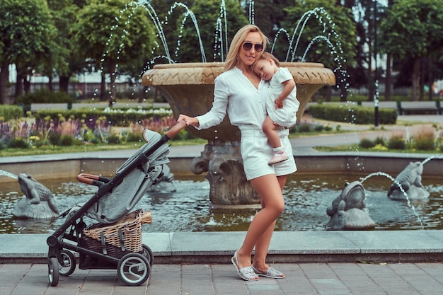 Heureuse femme blonde vêtue d'un chemisier blanc, d'un short et de lunettes de soleil tient sa petite fille sur les mains, posant près d'une fontaine dans le parc.