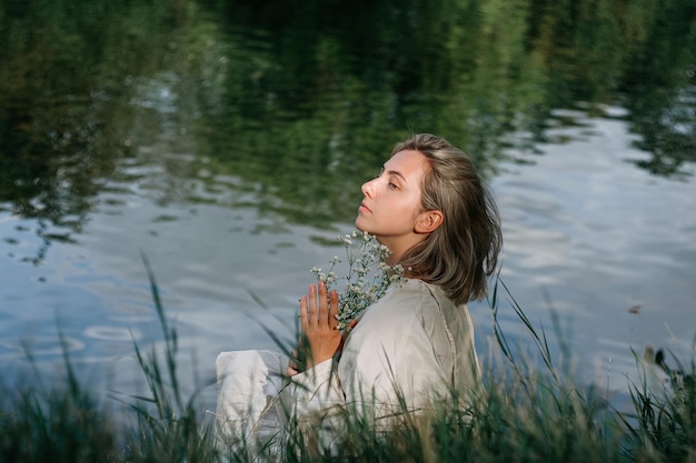 Heureuse femme blonde vêtue d'une robe vintage blanche assise et souriante près d'une rivière avec harmonie dans la nature.