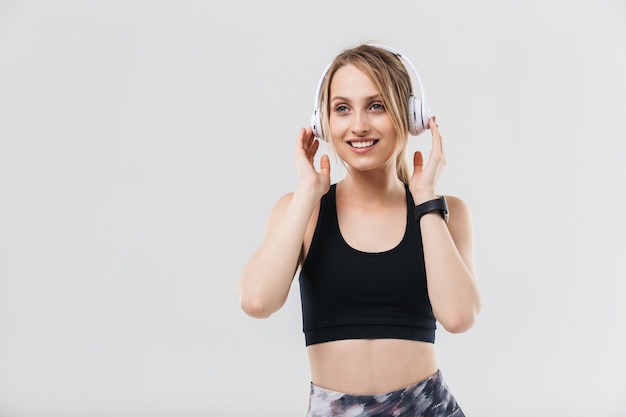 Heureuse femme blonde vêtue de vêtements de sport écoutant de la musique avec des écouteurs pendant l'entraînement dans une salle de sport isolée sur un mur blanc