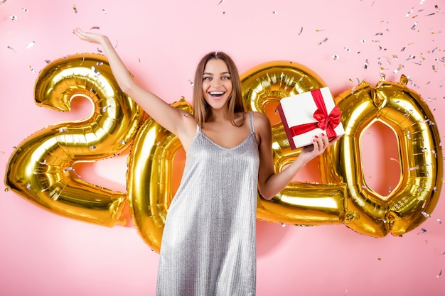 Heureuse femme avec une boîte-cadeau et des confettis d'argent et or 2020 ballons de nouvel an isolés sur rose