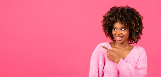 Photo heureuse femme bouclée afro-américaine du millénaire en tenue décontractée avec bretelles bouche ouverte pointer le doigt sur l'espace libre