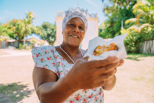 Heureuse femme brésilienne vêtue du costume traditionnel bahianais de la religion Umbanda, offrant des acarajÃ © - cuisine typique de Bahia - dans le centre historique de Porto Seguro en arrière-plan