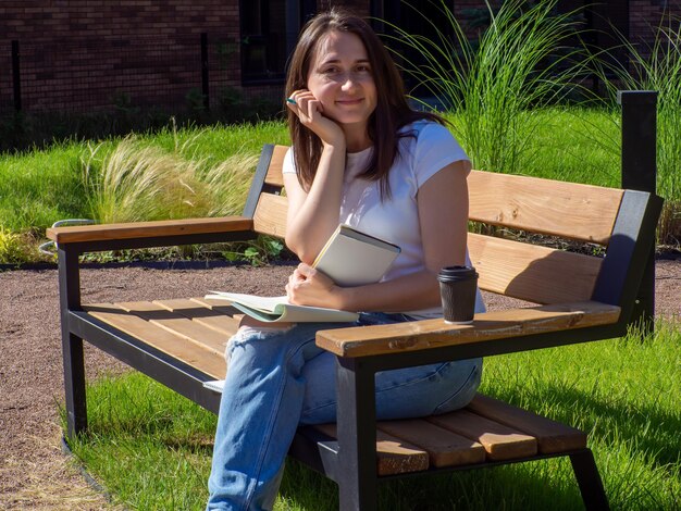 Heureuse femme brune assise sur un banc et lisant un livre dans le parc Éducation école et concept de personnes Étudiante étudiant sur un banc sur le campus Fille assise avec une tasse de café de livre et écrivant
