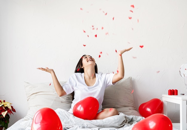 Heureuse femme brune assise dans le lit avec des ballons en forme de coeur rouge jetant des confettis en l'air