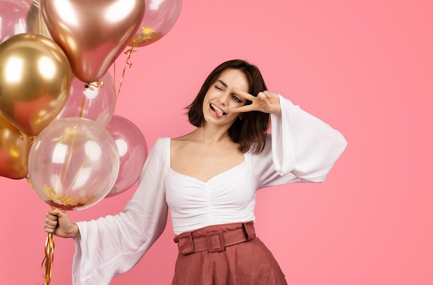 Heureuse femme brune caucasienne du millénaire avec de nombreux ballons gonflables faisant signe de paix