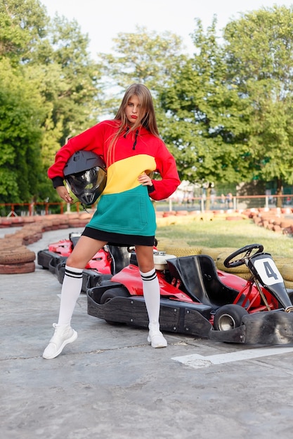 Heureuse femme brune gagnant la course de karting. Jeune fille blonde en costume de sport rouge posant en été sur une piste de karting