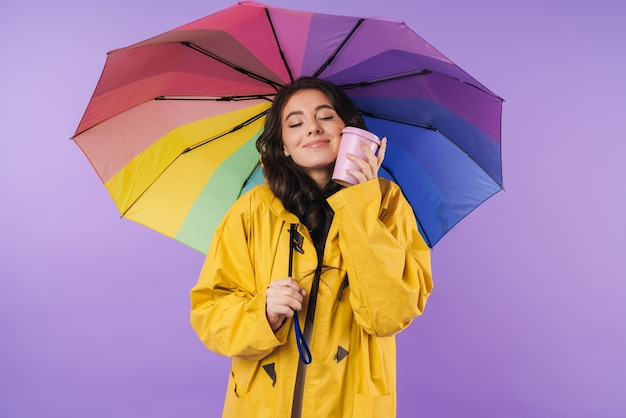 Heureuse femme brune joyeuse en imperméable jaune posant isolée sur un mur violet tenant un parapluie et du café.