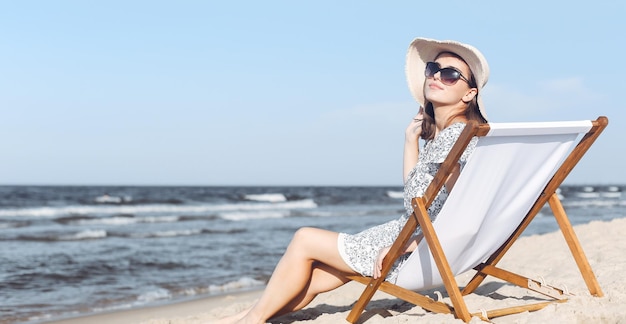 Heureuse femme brune portant des lunettes de soleil et un chapeau se reposant sur une chaise longue en bois sur la plage de l'océan.