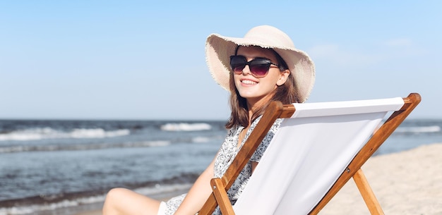 Heureuse femme brune portant des lunettes de soleil et un chapeau se reposant sur une chaise longue en bois à la plage de l'océan
