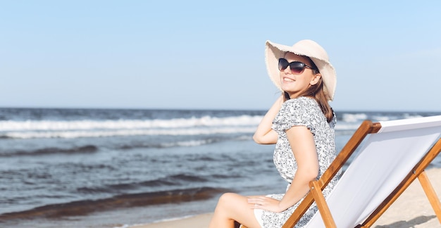 Heureuse femme brune portant des lunettes de soleil tout en vous relaxant sur une chaise longue en bois sur la plage de l'océan.