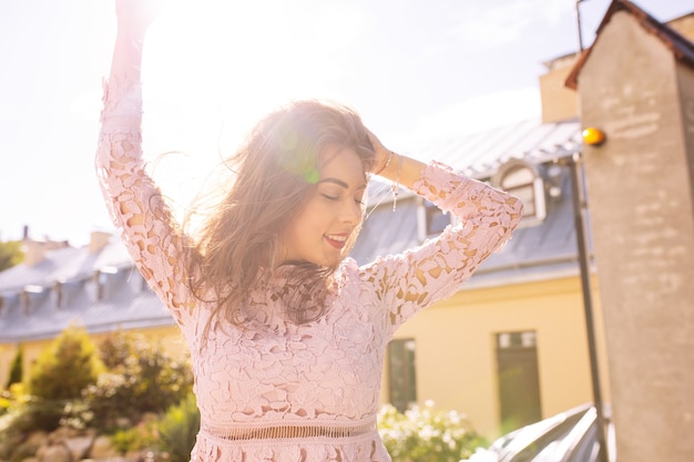 Heureuse femme brune en robe de dentelle rose posant dans la rue dans les rayons du soleil