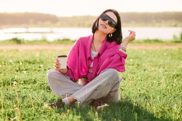 Heureuse femme brune en tenue décontractée assise sur la pelouse et se reposant dans le parc en journée ensoleillée