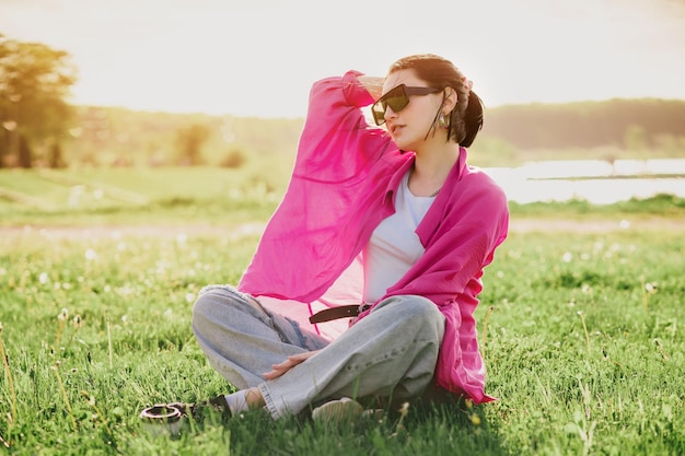 Heureuse femme brune en tenue décontractée assise sur la pelouse et se reposant dans le parc en journée ensoleillée