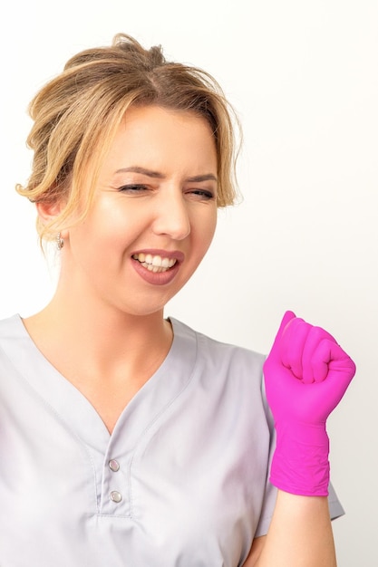 Heureuse femme caucasienne médecin portant des gants roses célèbre et lève les poings sur fond blanc