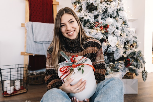 Heureuse femme caucasienne tenant grand cadeau de Noël assis sur le sol à la maison près de l'arbre de Noël.