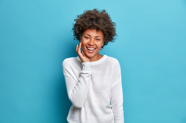 Heureuse femme charmante aux cheveux bouclés sourit largement touche le visage jouit d'une peau douce a la beauté naturelle exprime des émotions positives habillé cavalier incasuel isolé sur le mur bleu du studio