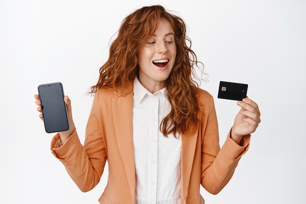 Heureuse femme chef de bureau souriante montrant une carte de crédit et un écran de téléphone portable recommandant une banque debout en costume sur fond blanc