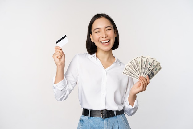 Heureuse femme coréenne tenant une carte de crédit et des dollars d'argent souriant et riant posant sur fond de studio blanc