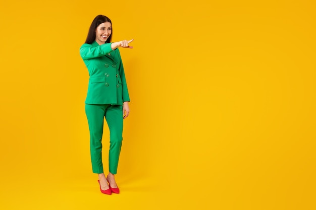 Heureuse femme élégante debout et pointant du doigt un espace vide isolé sur fond jaune bannière pleine longueur