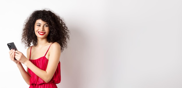 Heureuse femme élégante en robe rouge écrivant un message à l'aide d'un smartphone et souriant à la caméra debout aga