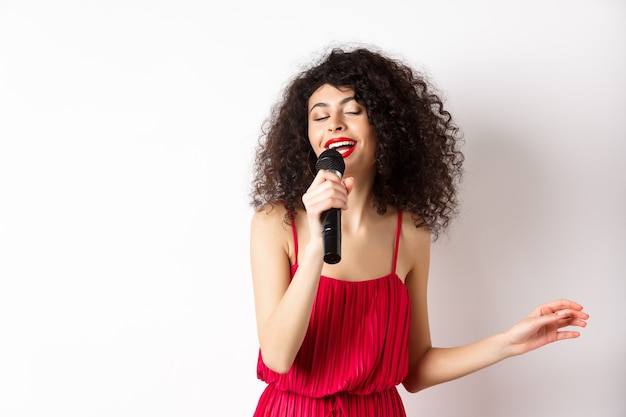 Heureuse femme élégante en robe rouge avec microphone, chant karaoké et souriant, debout sur fond blanc.