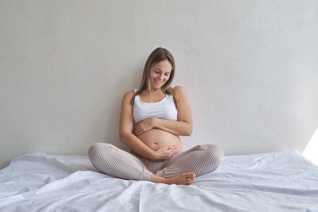 Heureuse femme enceinte assise sur le lit et touchant son ventre à la maison