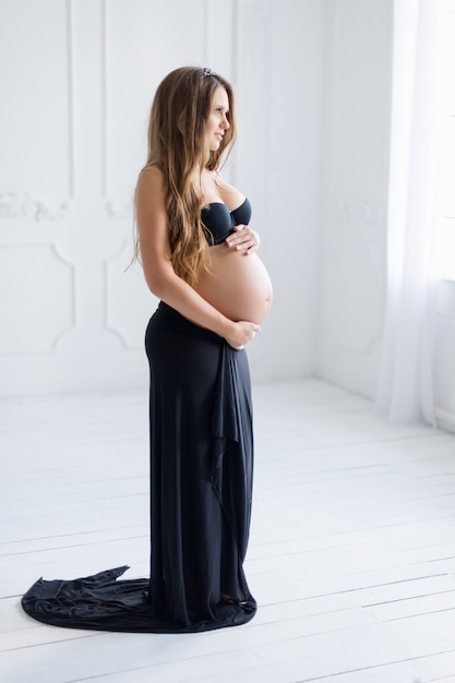 Heureuse femme enceinte aux cheveux longs dans un intérieur minimaliste blanc vêtu de lingerie noire.