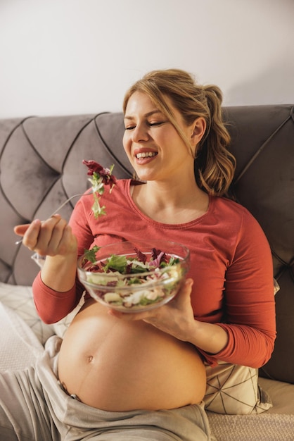 Heureuse femme enceinte mangeant une salade de légumes frais assise sur le lit dans sa chambre.