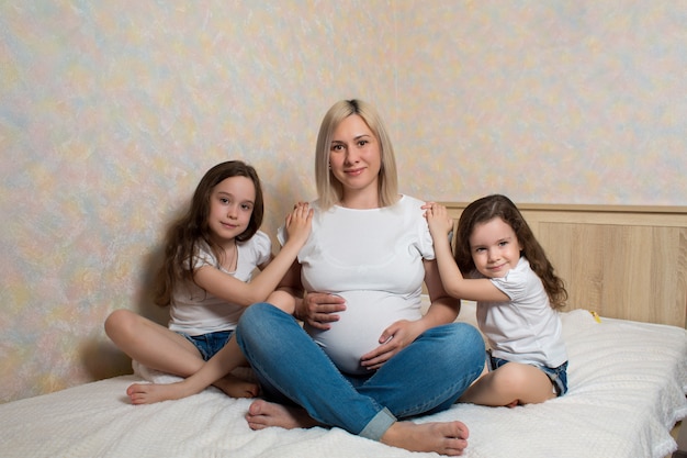 Photo heureuse femme enceinte avec ses filles, assise sur le lit à la maison. famille heureuse dans l'isolement. les enfants adorent leur mère