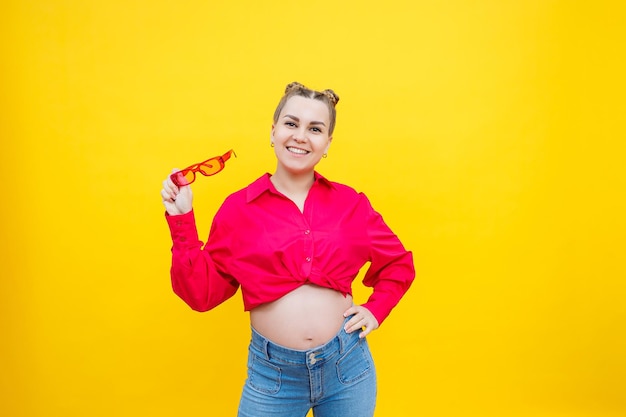 Heureuse femme enceinte avec un sourire sur son visage Joyeuse jolie femme enceinte dans une chemise rose et des lunettes roses sur fond jaune Jeune femme enceinte lumineuse