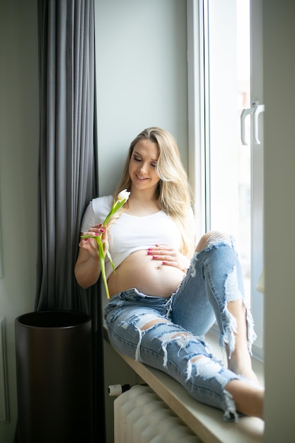 Heureuse femme enceinte tenir tulipe fraîche et toucher le ventre nu assis sur le rebord de la fenêtre photo verticale de la maison
